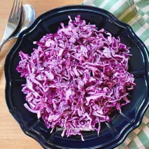 A bowl of red cabbage salad, on a wooden table, with a serving fork and spoon.